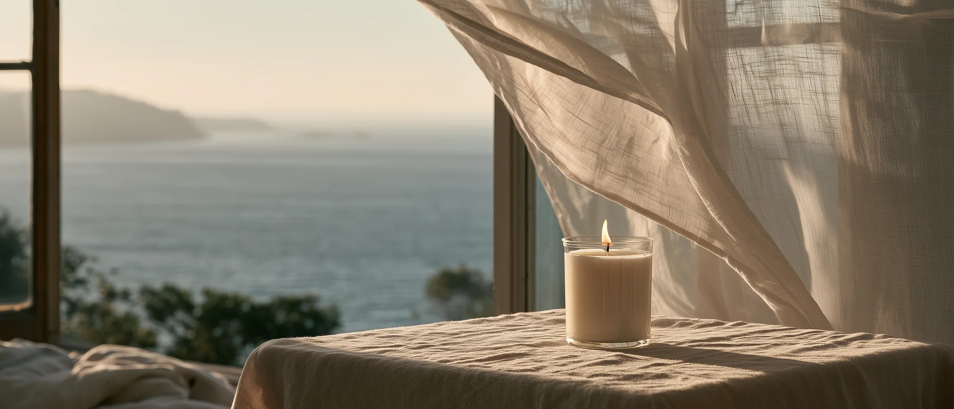 A lit soy candle on a linen-covered table by an open window, looking out over the sea